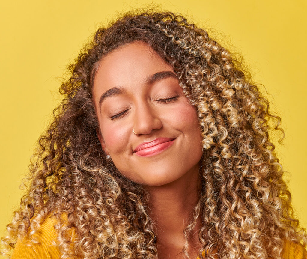 A young woman with curly hair with closed eyes and a big smile.