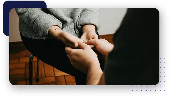 A seated individual with hands on knees, holding hands with another person for reassurance.